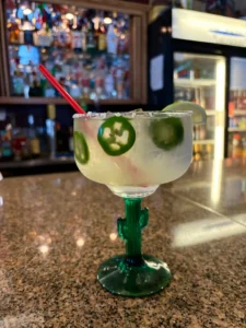 Refreshing margarita with a salted rim, garnished with a lime wheel and a slice of jalapeño pepper, served in a unique cactus-stemmed glass at a vibrant bar inside of El Palmar Mexican Restaurant in Carmichael, CA