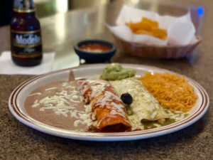 Plate of traditional Mexican enchiladas, one smothered in red sauce and the other in green sauce, both sprinkled with shredded cheese, alongside refried beans and Mexican rice, with a dollop of guacamole and a black olive. In the background is a bottle of negra Modelo beer and a basket of tortilla chips, suggesting a relaxed dining experience at El Palmar Mexican restaurant in Carmichael, CA