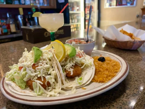 Fish tacos topped with fresh cabbage and a wedge of lime, paired with Spanish rice and a side of salsa, with a frozen margarita and a basket of tortilla chips in the background, ready to be enjoyed at El Palmar Mexican Restaurant in Carmichael, CA