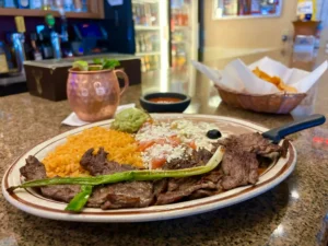 Grilled carne asada strips with roasted green onions on a plate alongside Mexican rice, refried beans topped with cheese, fresh guacamole, and pico de gallo, with a background featuring a copper Moscow mule mug and a basket of tortilla chips, at El Palmar Mexican restaurant Carmichael, CA